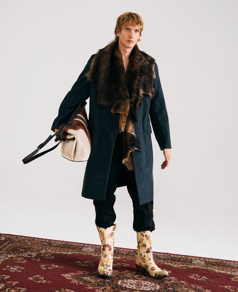 A male model poses indoors wearing a long coat with a fur collar, black pants, floral boots, and carrying a striped duffel bag.
