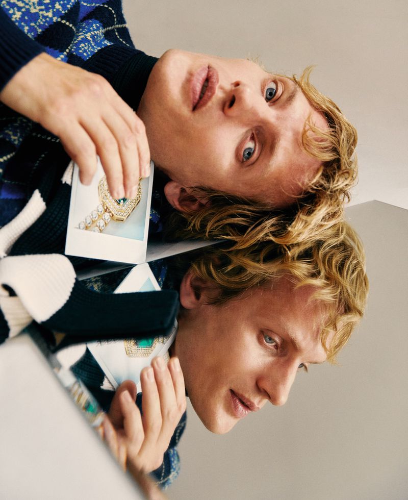 Young adult poses with mirrored reflection, holding a photo of a large diamond ring while wearing a patterned sweater.