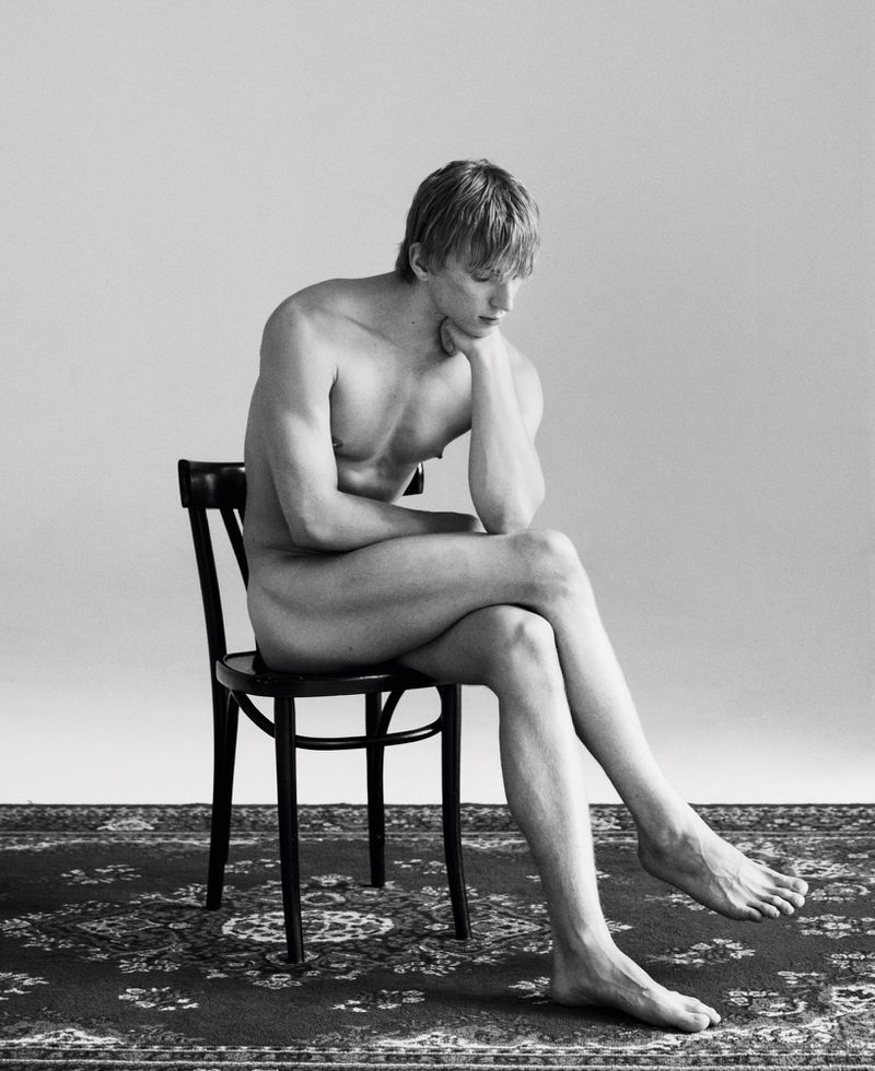 A nude male with blond hair sits on a black chair, legs crossed, in a black and white photo against a light grey backdrop.