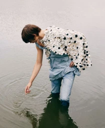 Model wading in calm water wearing a textured disc-embellished cape, blue layered tunic, and denim jeans, touching the surface.
