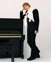 Male model in a black suit with a white ruffled scarf leaning against a black upright piano against a white seamless background.
