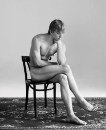 A nude young man with blond hair sits pensively on a dark wooden chair atop an ornate patterned rug against a neutral studio backdrop.