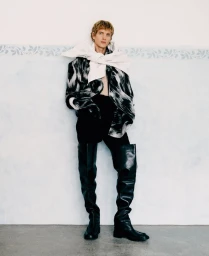Model wearing a black and white patterned jacket with thigh-high black boots, leaning against a pale blue wall.