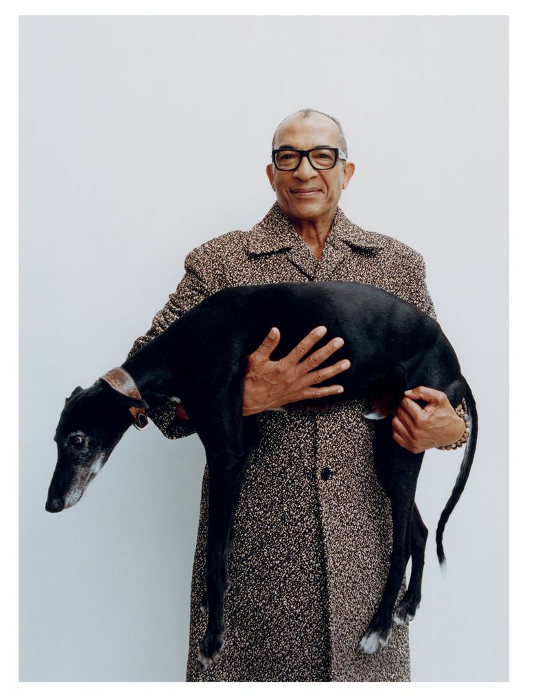 Elderly African-American man in patterned coat holds a black greyhound dog against a simple white background in a portrait shot.