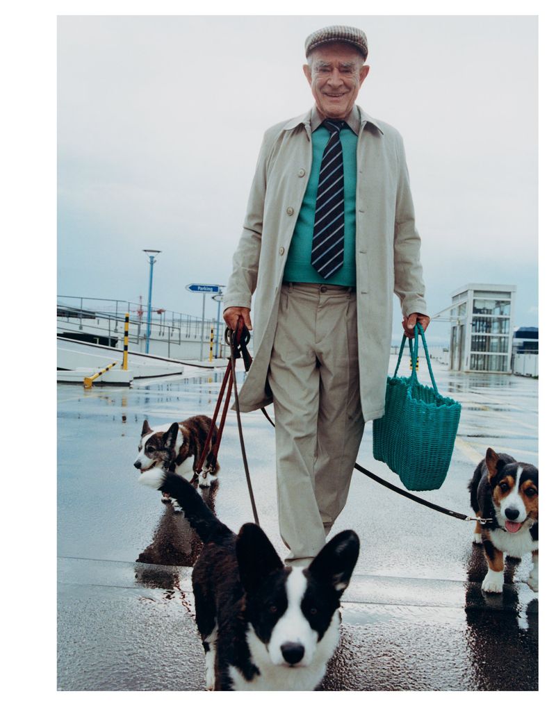 Elderly man walking three corgis outdoors on a wet day, carrying a green woven bag. He is wearing a flat cap and a tan trench coat.