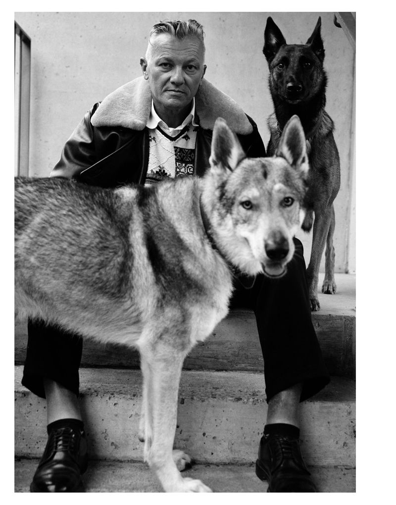 Black and white portrait of a man sitting on stairs with two dogs, one a German Shepherd, wearing a jacket and patterned shirt.
