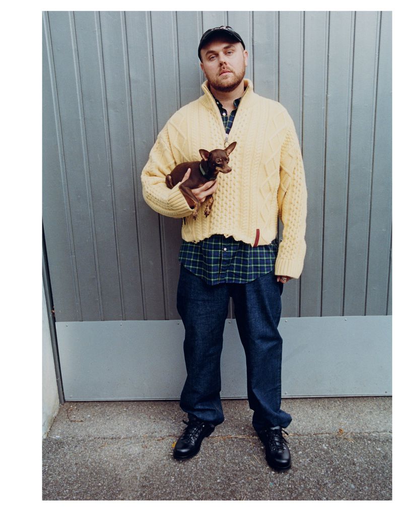 An adult male stands with a dog outside against a metal wall, wearing a yellow sweater, dark jeans and shoes and a baseball cap.