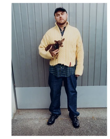 An adult male stands with a dog outside against a metal wall, wearing a yellow sweater, dark jeans and shoes and a baseball cap.