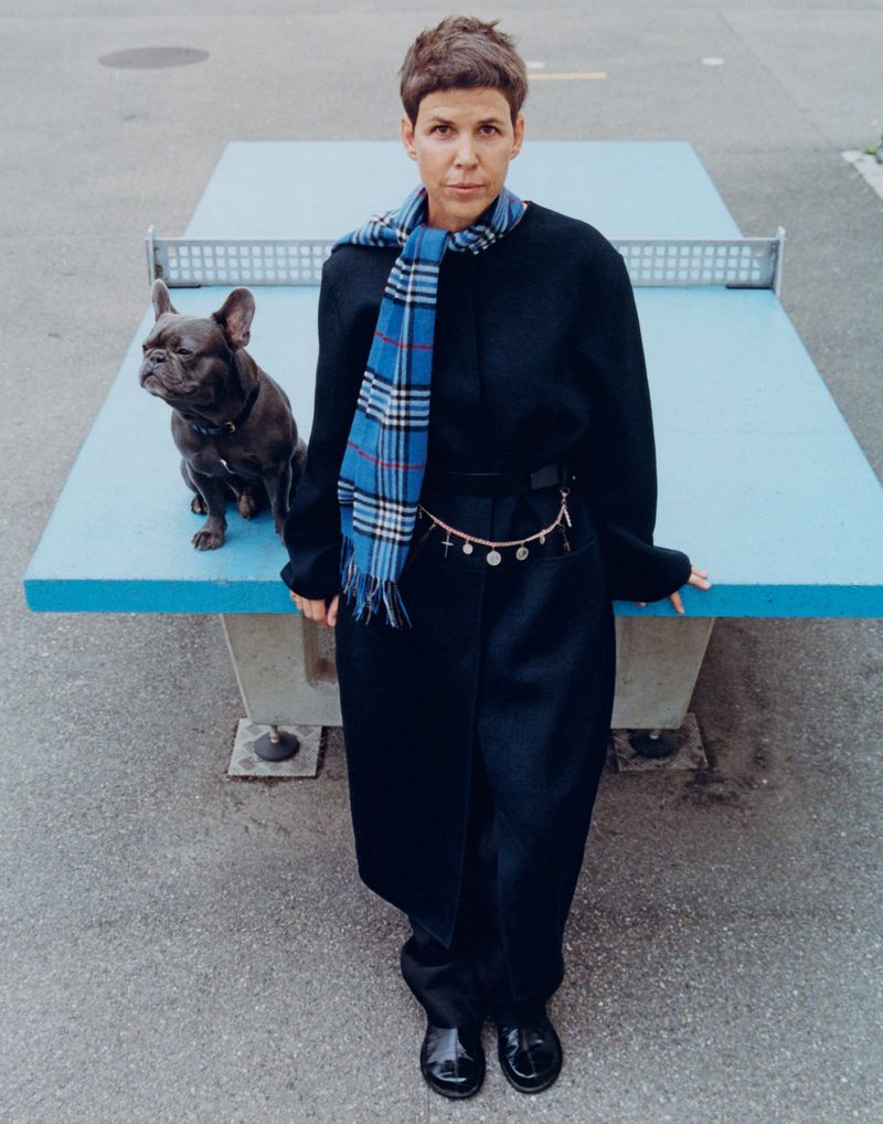 Woman in a black coat and blue scarf poses with a French bulldog next to a blue ping pong table on an asphalt surface.