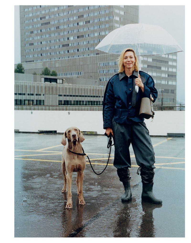 A woman in a blue jacket holds an umbrella and a dog leash on a rainy rooftop with a city building in the background.