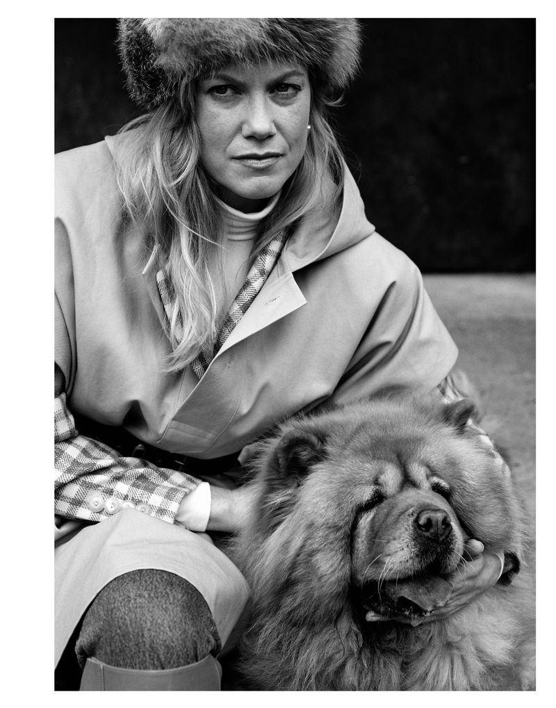 Black and white portrait of a blonde adult woman in a fur hat, holding a fluffy Chow Chow dog with her hands, posing outdoors.