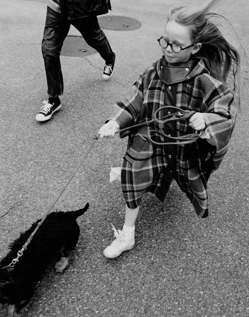 Child with glasses and plaid coat runs with dog on leash, person walks in background, captured in black and white, urban environment.