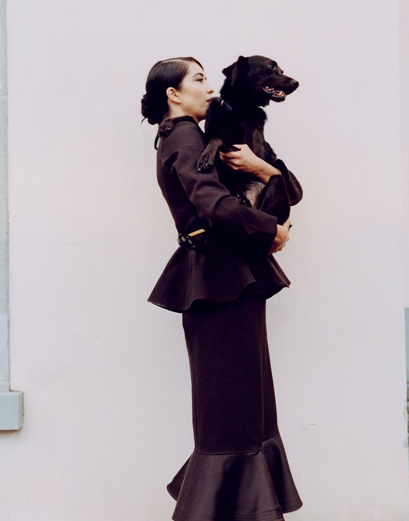 An adult woman in a dark two-piece suit poses outdoors while holding a dark dog in her arms against a light-colored wall.
