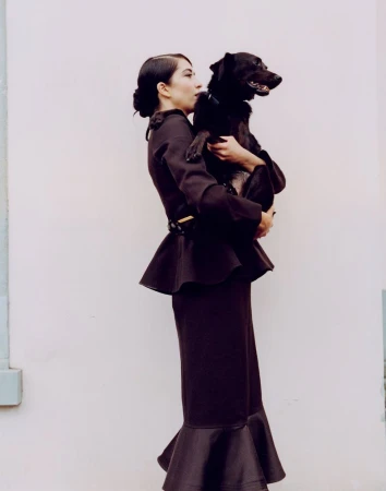 An adult woman in a dark two-piece suit poses outdoors while holding a dark dog in her arms against a light-colored wall.