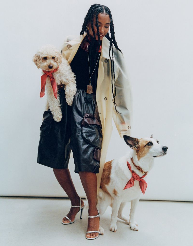 A young Black woman poses indoors with a white poodle and a white/tan Jack Russell Terrier, both dogs wearing matching red bandanas.