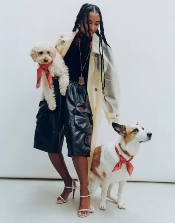 A young Black woman poses indoors with a white poodle and a white/tan Jack Russell Terrier, both dogs wearing matching red bandanas.