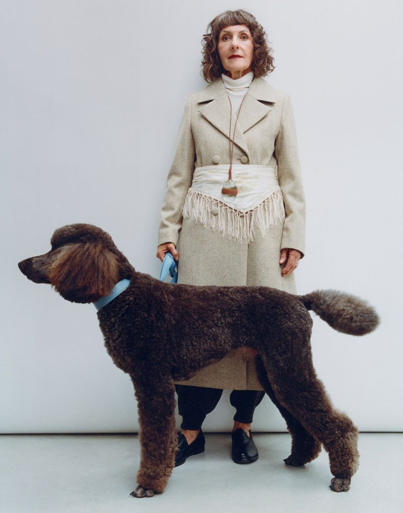 Elegant elderly woman in beige coat stands next to a large brown poodle with a blue collar against a neutral background in a portrait.