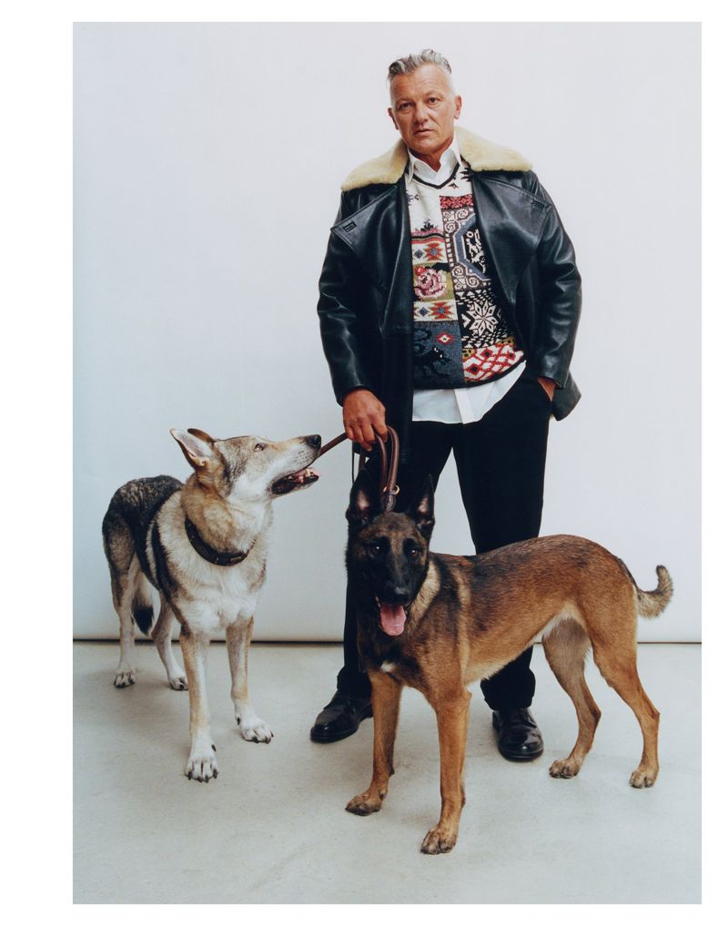 Man poses with two dogs in a studio. He wears a black leather jacket and the dogs wear collars with light grey background.