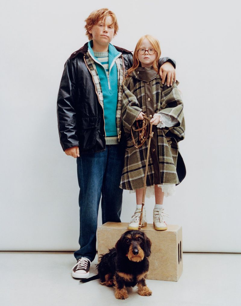 Two children and a dachshund dog pose in a studio setting. The girl stands on a beige block, wearing a plaid coat, while the boy wears a black jacket.