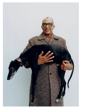 An older man wearing glasses and a textured patterned coat holding a sleek black dog against a white background.