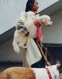Woman in layered cream coat holding a fluffy white poodle wearing a pink scarf outdoors