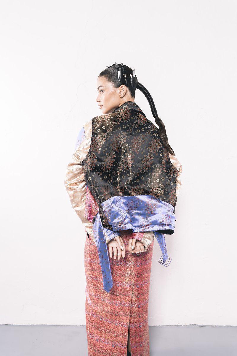 A woman with dark hair styled in a thick braid and wearing a patterned jacket and long skirt poses in front of a white wall.