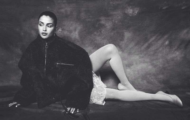 Black and white portrait of a woman reclining, wearing a dark jacket, light skirt and tights against a mottled backdrop.