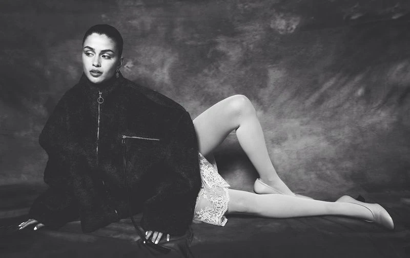 Black and white portrait of a woman reclining, wearing a dark jacket, light skirt and tights against a mottled backdrop.