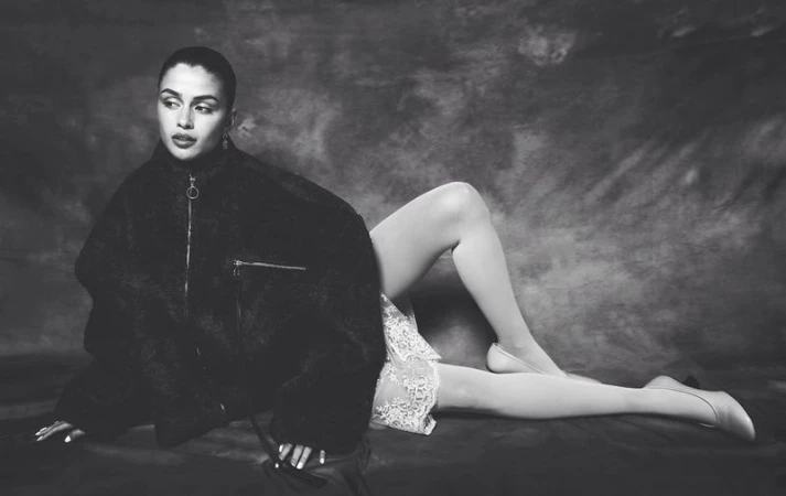 Black and white portrait of a woman reclining, wearing a dark jacket, light skirt and tights against a mottled backdrop.