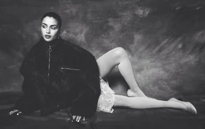 Monochrome editorial portrait of a woman reclining in an oversized black jacket, lace slip dress, and heels against a textured backdrop.