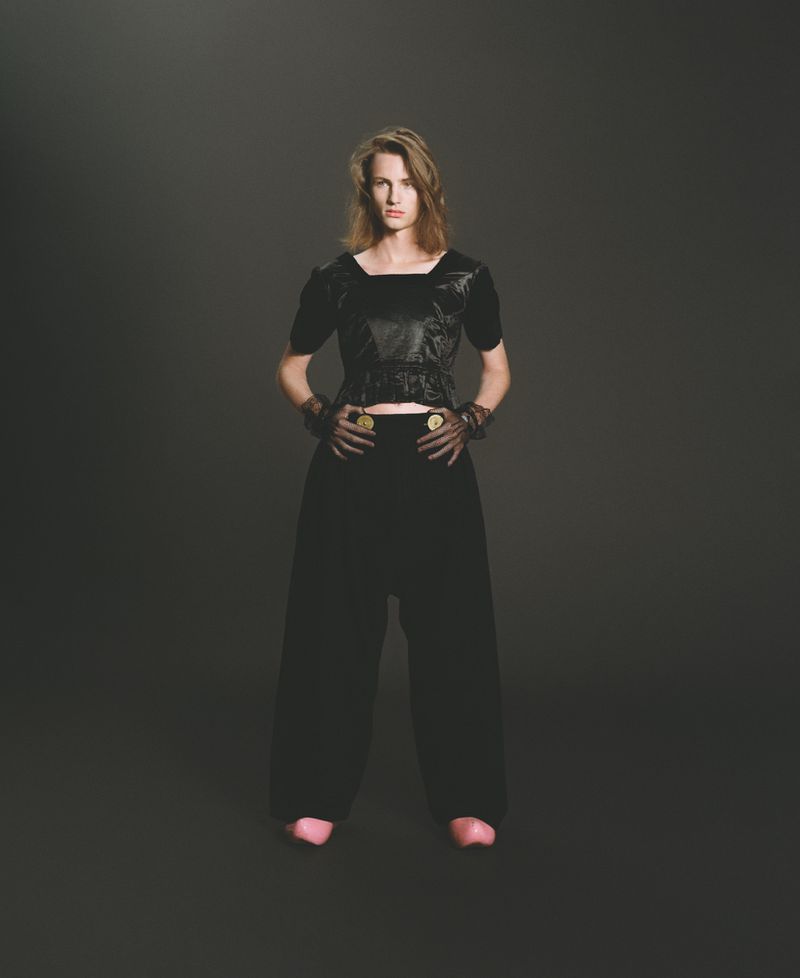 Full shot of a fair-skinned adult woman in a black textured top and loose black pants posing against a dark gray background.