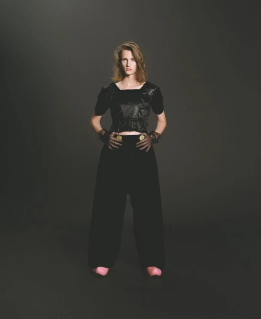Full shot of a fair-skinned adult woman in a black textured top and loose black pants posing against a dark gray background.