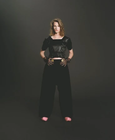 Full shot of a fair-skinned adult woman in a black textured top and loose black pants posing against a dark gray background.