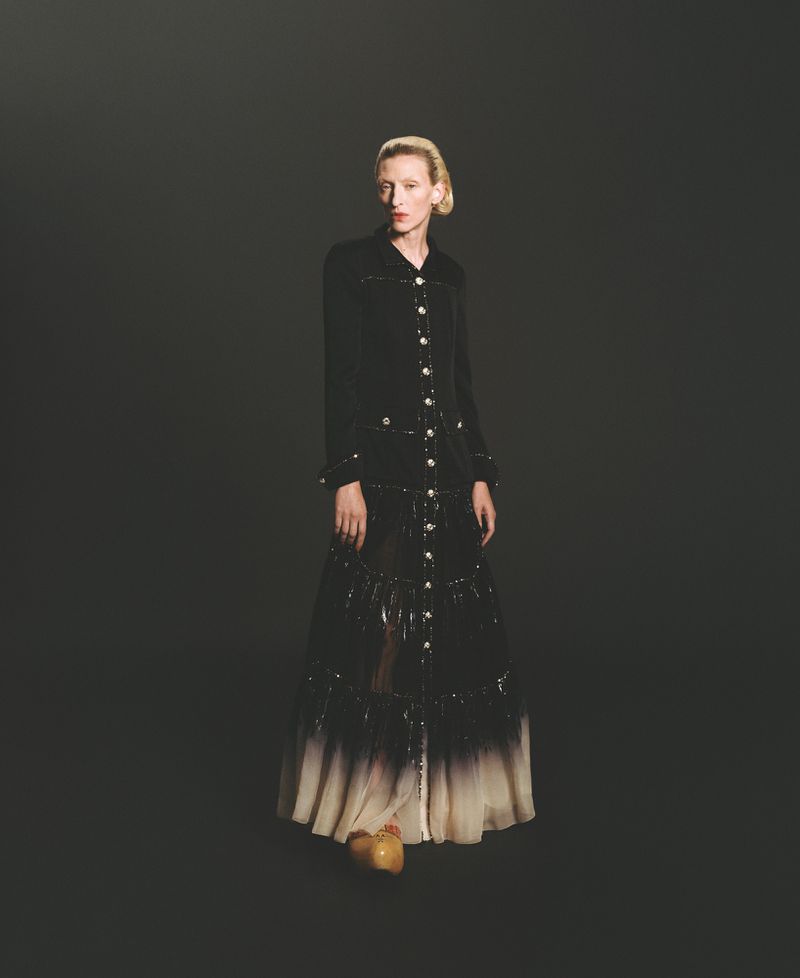 A fair-skinned woman with blonde hair stands in a black coat dress with button details and tan platform shoes, against a dark background.