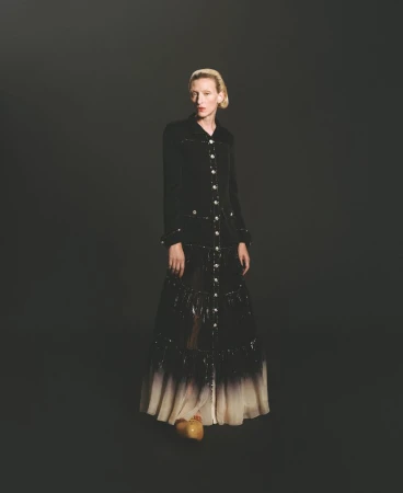 A fair-skinned woman with blonde hair stands in a black coat dress with button details and tan platform shoes, against a dark background.