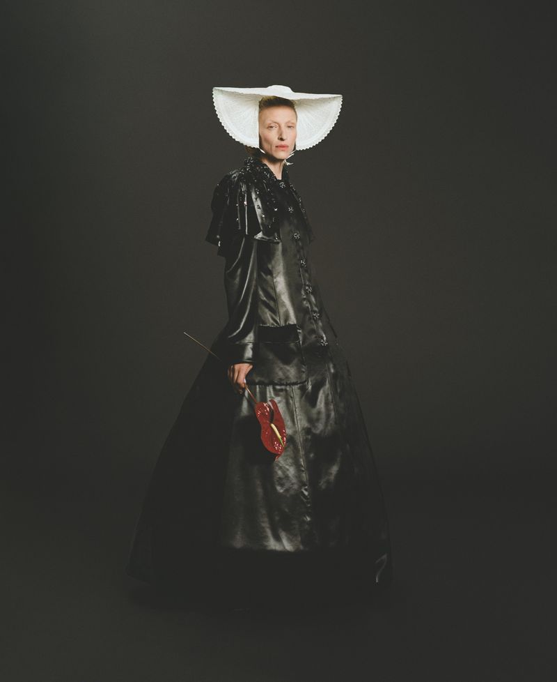 Woman in dramatic black dress and white bonnet holds a red violin in a studio setting with dark background, fashion editorial.