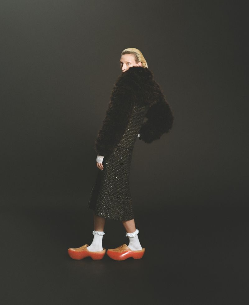 Fashionable adult female model in a patterned midi skirt suit with fur accents and orange platform clogs against a gray backdrop.