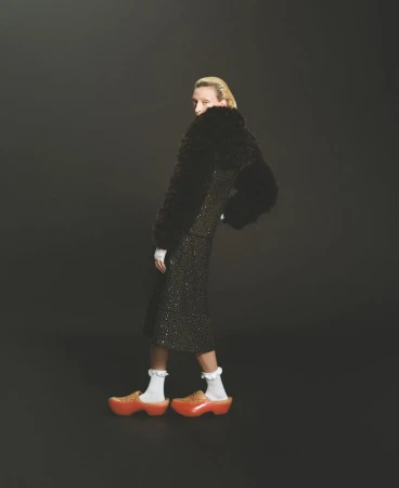 Fashionable adult female model in a patterned midi skirt suit with fur accents and orange platform clogs against a gray backdrop.