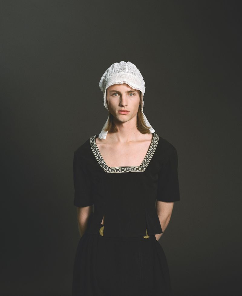 Portrait of a person with light skin, wearing a black dress with a white lace bonnet, set against a dark grey studio backdrop.