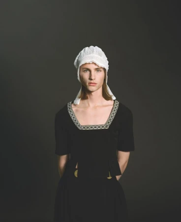 Portrait of a person with light skin, wearing a black dress with a white lace bonnet, set against a dark grey studio backdrop.