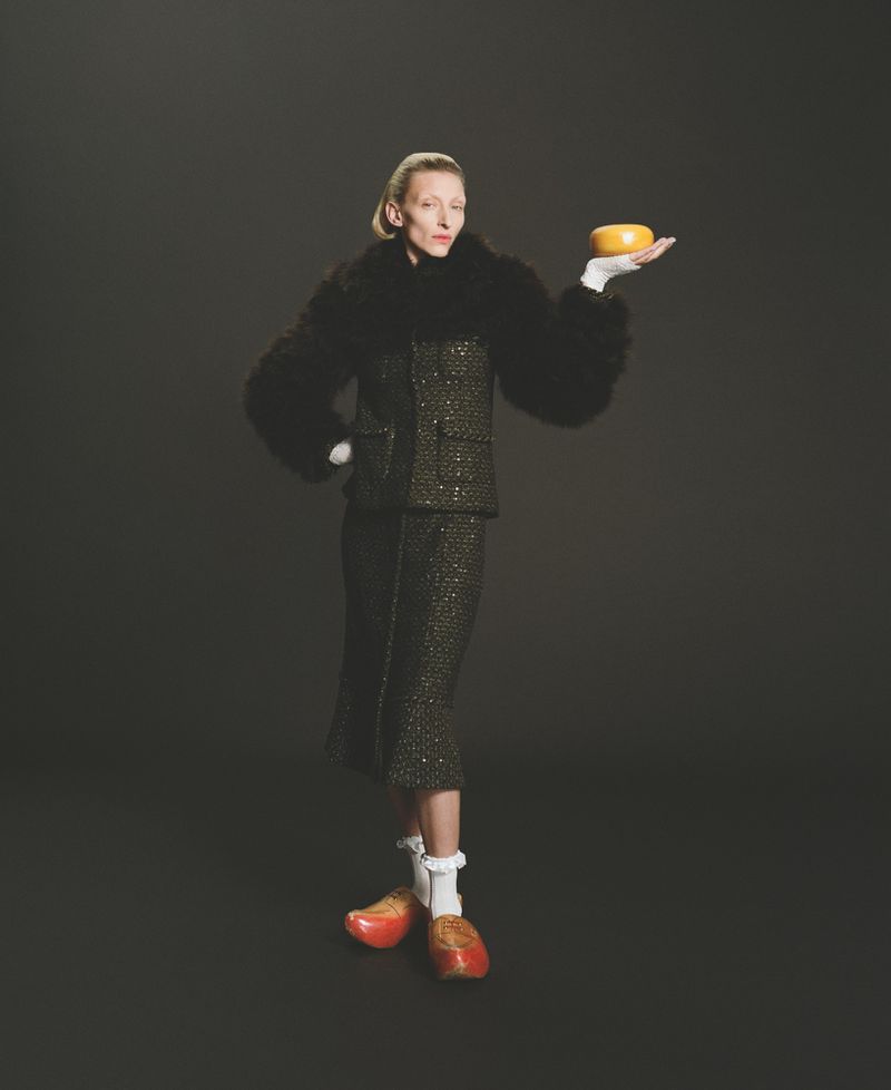 Woman in tweed skirt suit holds orange object in photo studio against dark backdrop, wearing vintage wooden shoes and white gloves.