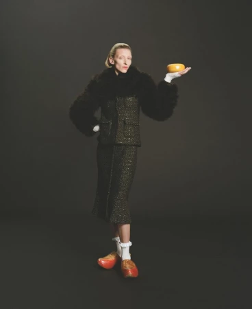 Woman in tweed skirt suit holds orange object in photo studio against dark backdrop, wearing vintage wooden shoes and white gloves.