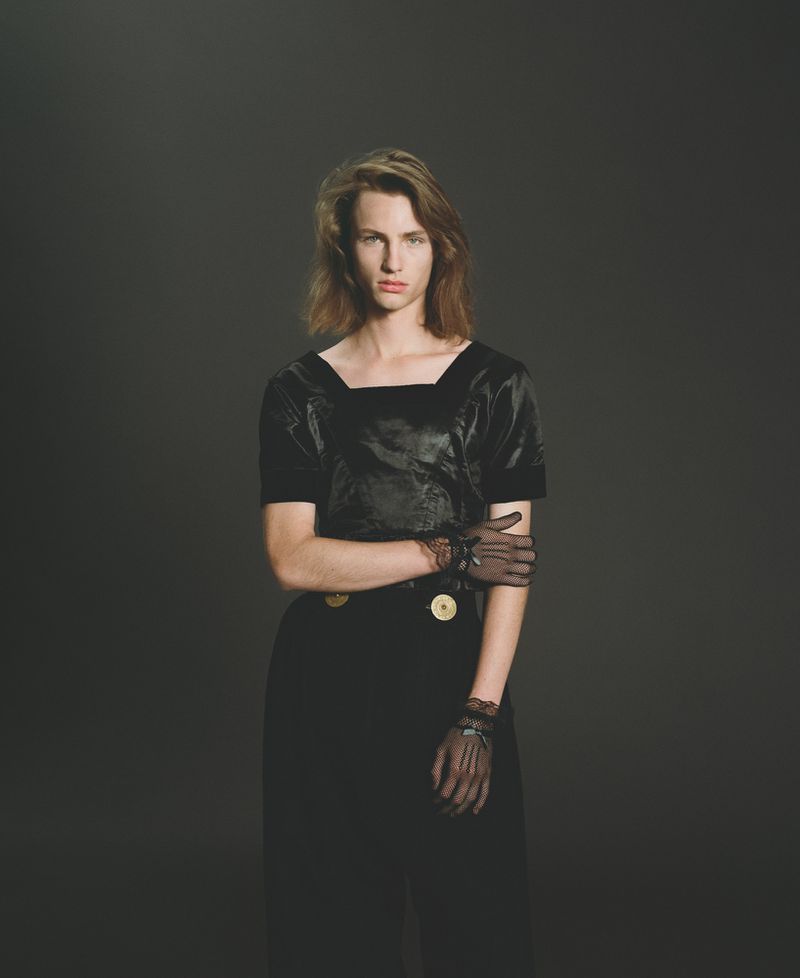 Studio portrait of an androgynous person in black dress and sheer gloves posing against a dark backdrop with simple, elegant studio lighting.