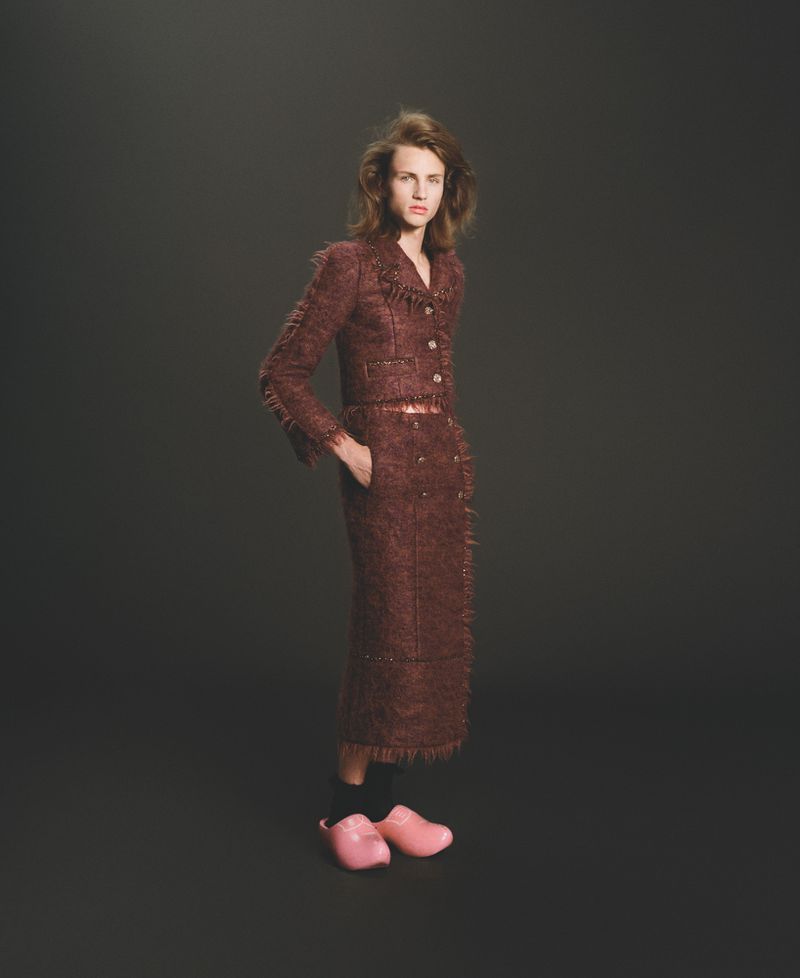 Model Maggie Maurer poses in a fringed brown tweed skirt suit and pink clogs against a dark studio background.