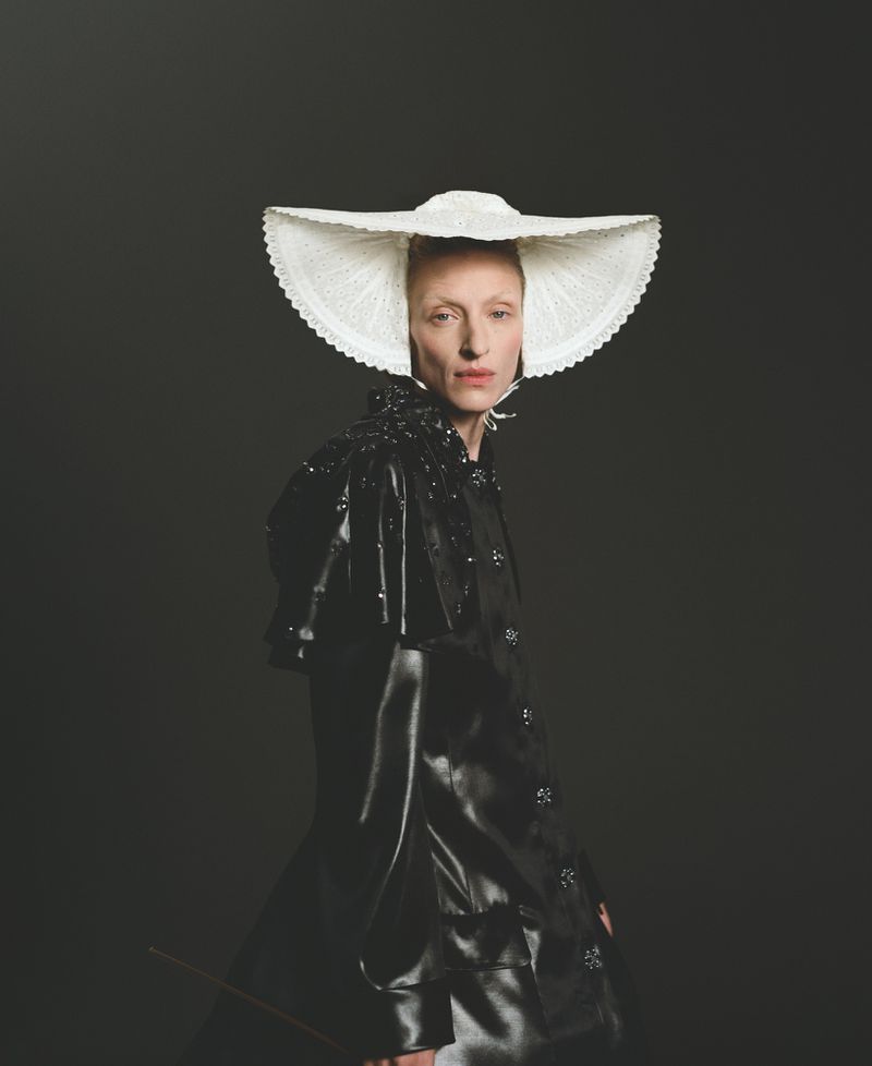 An adult female poses in a glossy black coat and large white lace hat against a dark background in a fashion portrait.