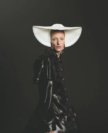 An adult female poses in a glossy black coat and large white lace hat against a dark background in a fashion portrait.