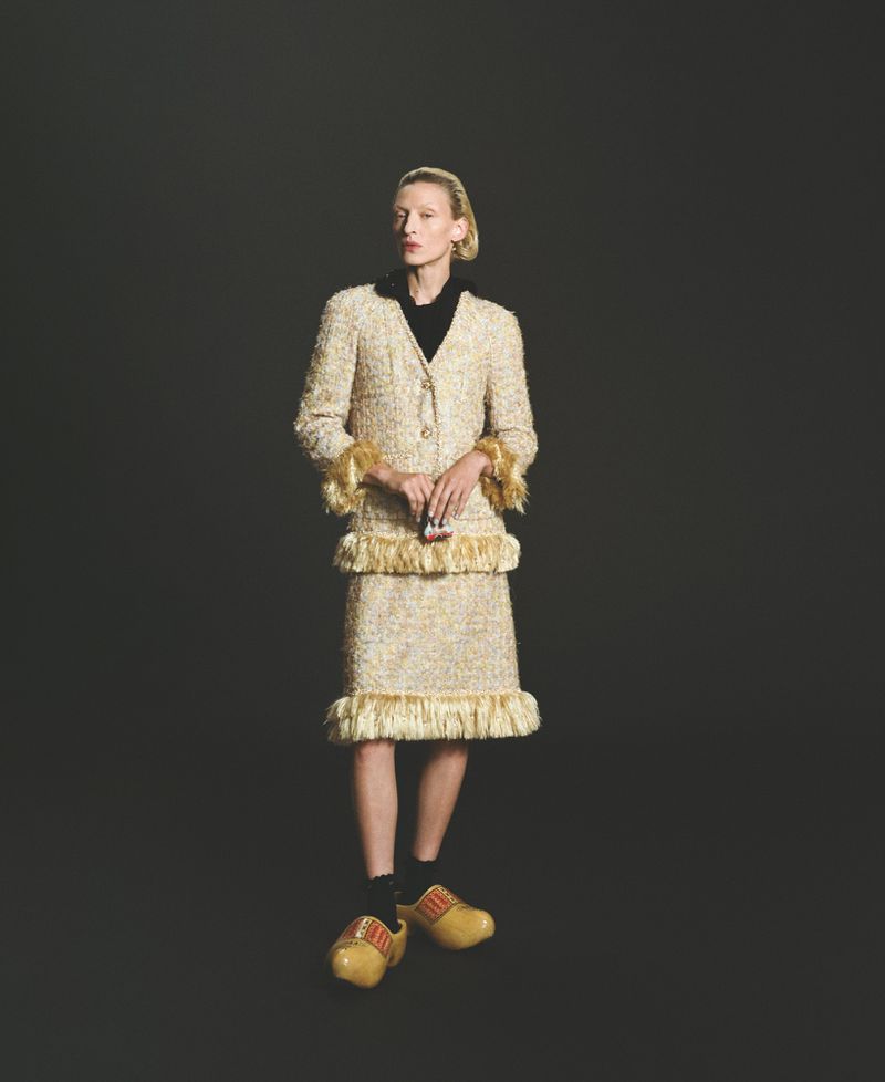 Full-length shot of a slender blonde model posing in a tweed skirt suit with fringe and yellow clogs against a dark grey backdrop.