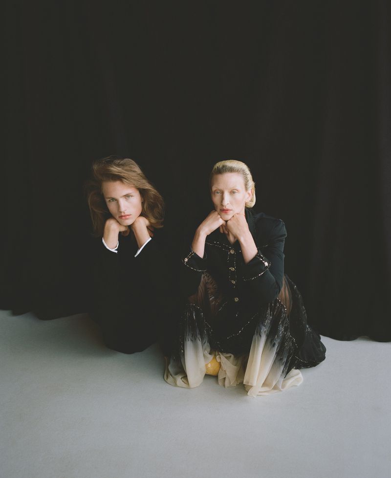 Two fashion models pose kneeling on a gray floor against a black background in a studio. One wears a black suit, the other a dress.