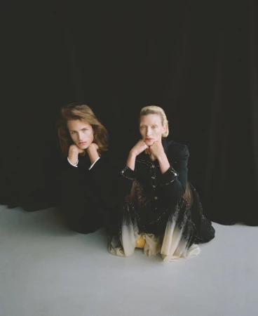 Two fashion models pose kneeling on a gray floor against a black background in a studio. One wears a black suit, the other a dress.