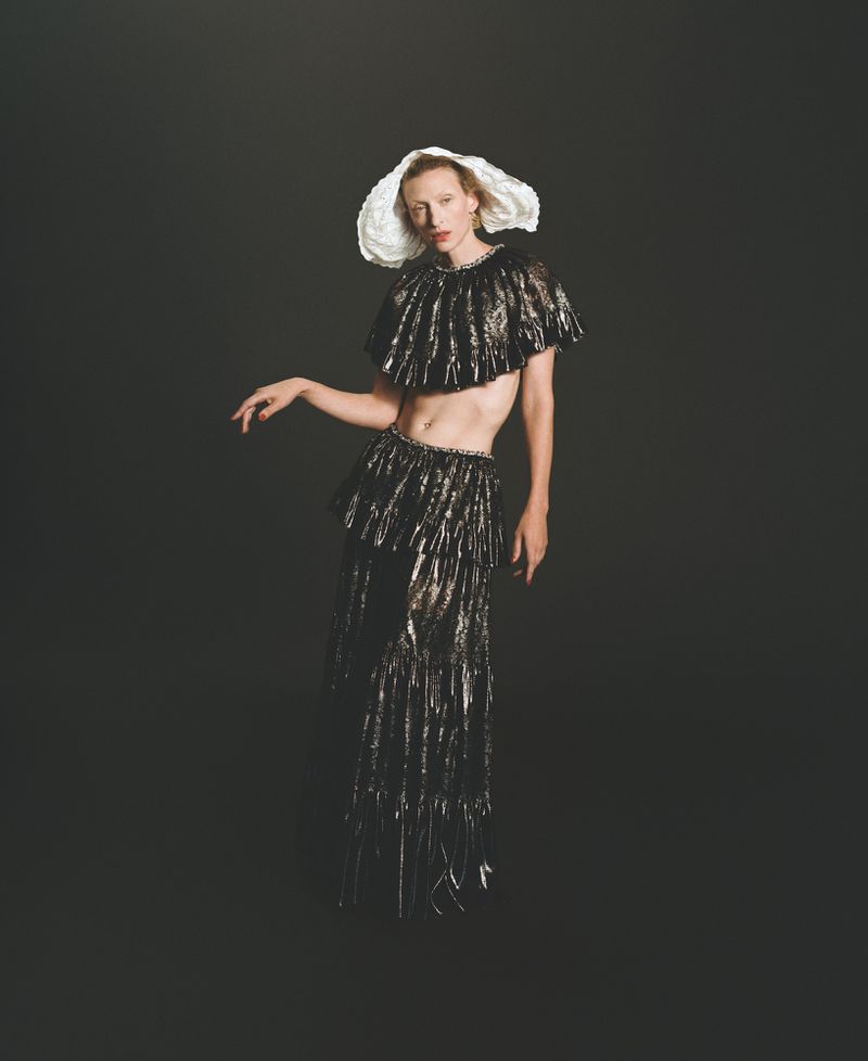 Elegant individual poses in a metallic fringe outfit and white hat on a dark backdrop in a studio fashion portrait, showcasing high fashion.
